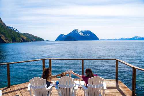 Three friends toast with glasses on a deck overlooking a serene lake and snow-capped mountains under a clear blue sky.