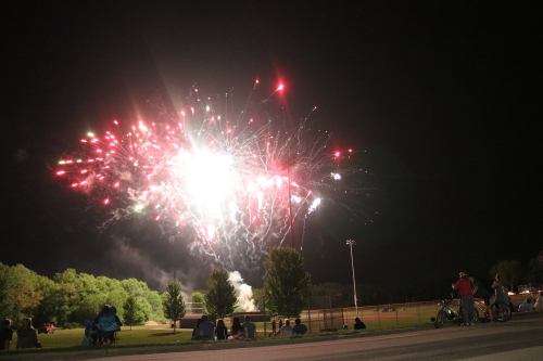Colorful fireworks illuminate the night sky over a park, with spectators sitting on the ground and enjoying the display.