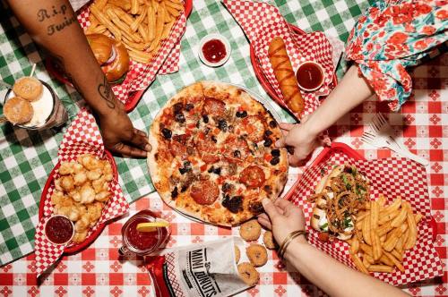 A table filled with pizza, burgers, fries, and drinks, with hands reaching for the pizza. Red and white checkered tablecloth.