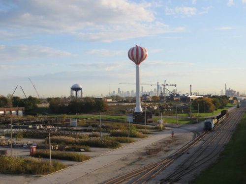 A landscape featuring water towers, industrial structures, and train tracks under a blue sky with scattered clouds.