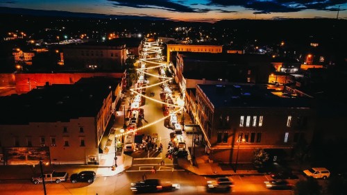 Aerial view of a city street illuminated by string lights at dusk, with cars parked along the sides.