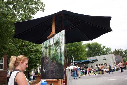 A woman paints under a large umbrella at an outdoor event, with a crowd and buildings in the background.