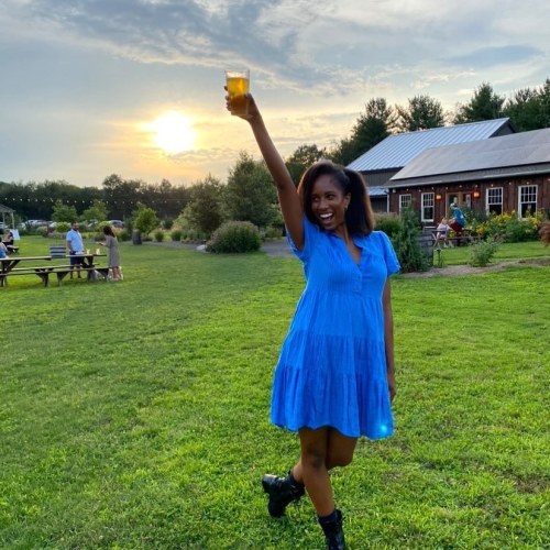 A person in a blue dress raises a drink in celebration, with a sunset and outdoor gathering in the background.