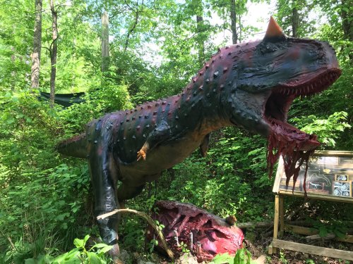 A large, colorful dinosaur statue with a fierce expression, surrounded by lush greenery and a display board nearby.