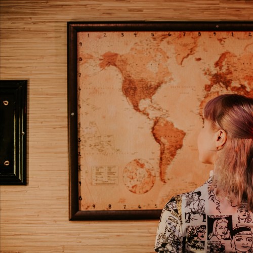 A person with light purple hair stands in front of a vintage world map, wearing a patterned shirt.