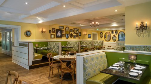 Cozy restaurant interior with green booths, wooden tables, and decorative mirrors on yellow walls. Warm, inviting atmosphere.