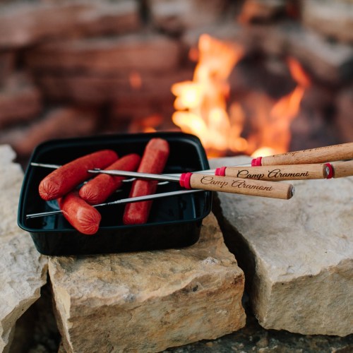 A black tray with hot dogs and roasting sticks sits near a campfire and stone backdrop.