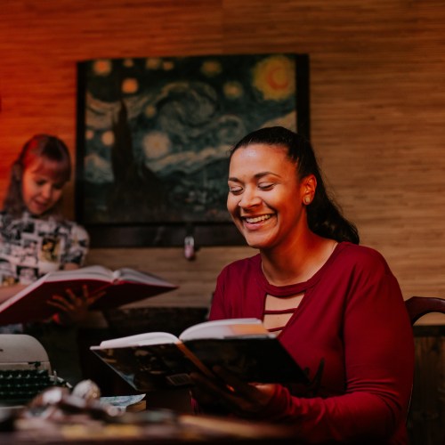 Two women in a cozy setting, one reading a book and smiling, while the other looks at a book in the background.