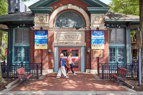 Two women walk past a historic market building with banners for a spelling bee event. Benches line the walkway.