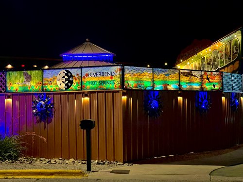 Colorful stained glass panels illuminate the entrance of Riverbend Hot Springs at night, surrounded by decorative lights.