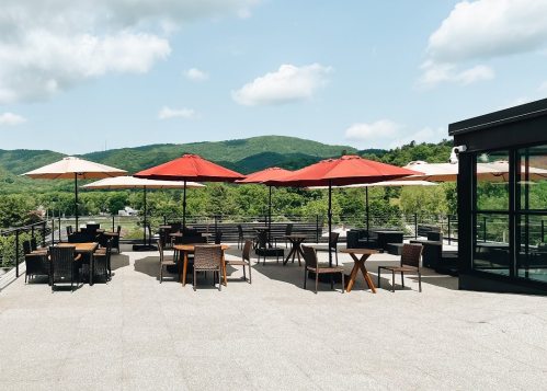 Outdoor seating area with wooden tables and umbrellas, surrounded by green hills and a clear blue sky.