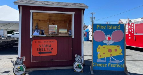 A small red storm shelter booth next to a colorful sign for the Pine Island Cheese Festival, featuring cartoon mice.