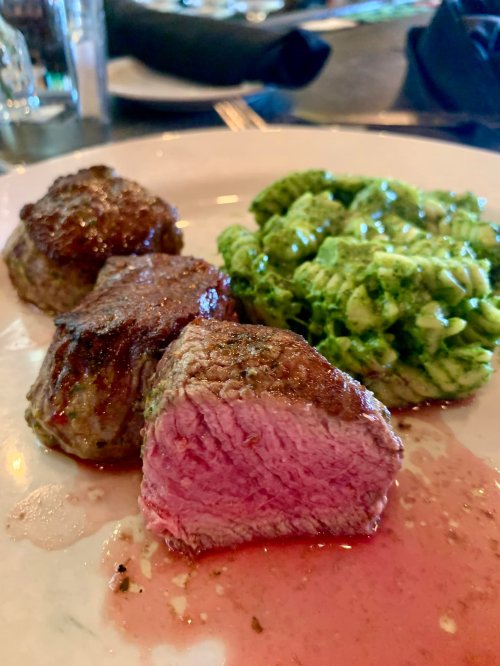 A plate featuring three cooked beef medallions and a serving of green pesto pasta. One medallion is sliced open, revealing pink meat.