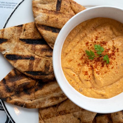 A bowl of hummus garnished with paprika and herbs, surrounded by grilled pita bread on a plate.