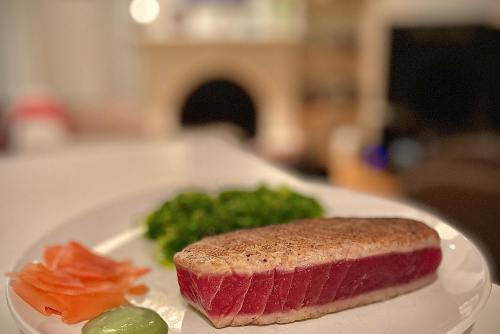 A plate with seared tuna steak, sliced salmon, green vegetables, and a dollop of wasabi. Soft background blur.