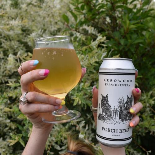 A hand holds a glass of light beer and a can labeled "Porch Beer" from Arrowood Farm Brewery, surrounded by greenery.