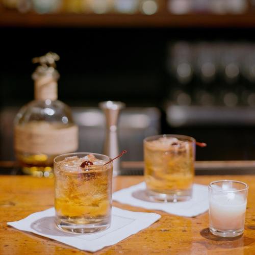 Two glasses of cocktails with cherries on a bar, alongside a bottle and a small candle in the background.