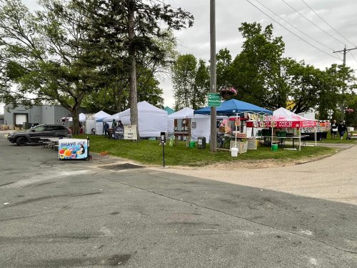 A view of a market area with white tents, food stalls, and a sign for shaved ice, surrounded by trees.