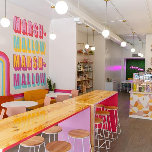 Bright, colorful interior of a café with a "Marshmallow" wall, wooden bar, and playful decor. Cozy seating area visible.