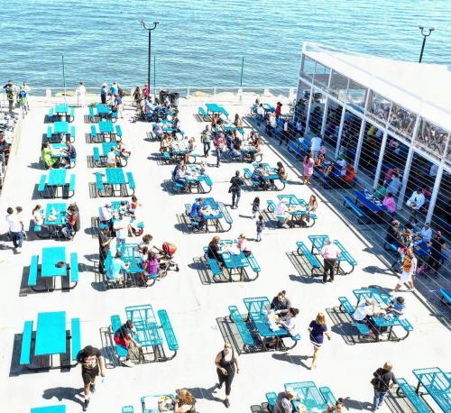 A busy outdoor dining area with turquoise tables, people enjoying meals by the water on a sunny day.