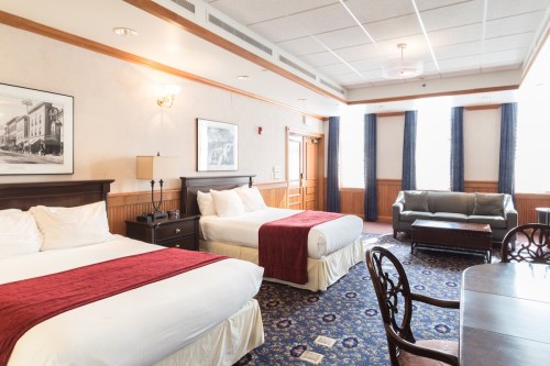 A spacious hotel room with two beds, a seating area, and large windows, featuring classic decor and warm colors.