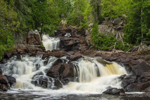 A serene waterfall cascades over rocky terrain, surrounded by lush green trees and a tranquil river.