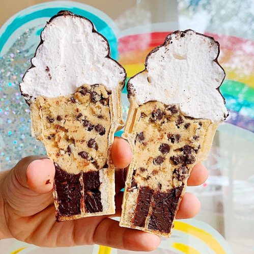 A hand holding two ice cream cone-shaped desserts filled with cookie dough and topped with whipped cream.