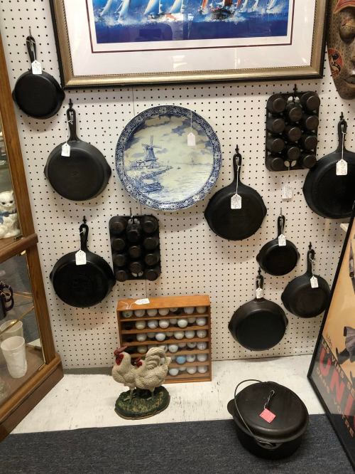 A display of various cast iron pans, a decorative plate, and a wooden egg holder, with a ceramic chicken in front.