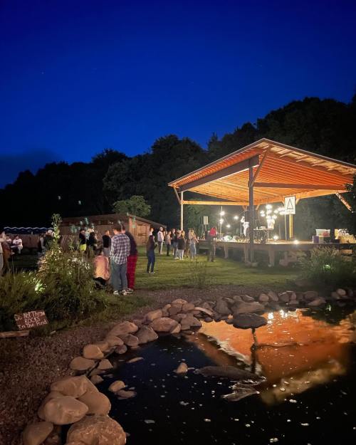 A lively outdoor event at night, with people gathered under a lit pavilion beside a pond reflecting the lights.
