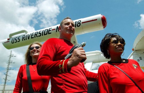 Three people in red shirts pose in front of a large model of a spaceship, smiling against a blue sky.