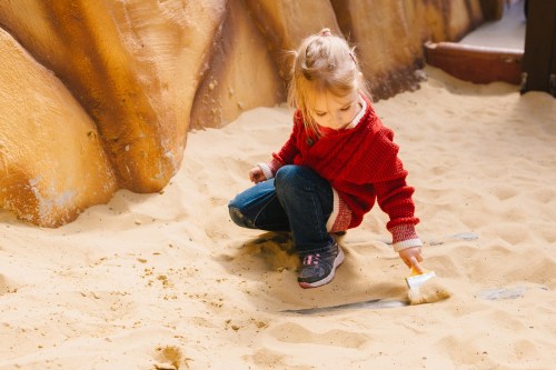 A young girl in a red sweater brushes sand with a small broom in a sandy play area.