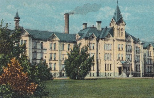 Historic building with a tower, surrounded by trees and a grassy area, featuring a smokestack in the background.