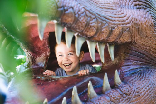A smiling child peeks out from inside a large dinosaur mouth, surrounded by greenery.