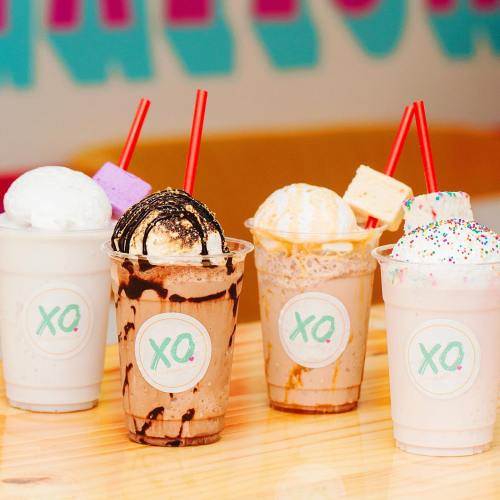 Four colorful milkshakes in clear cups with whipped cream, straws, and toppings, displayed on a wooden table.