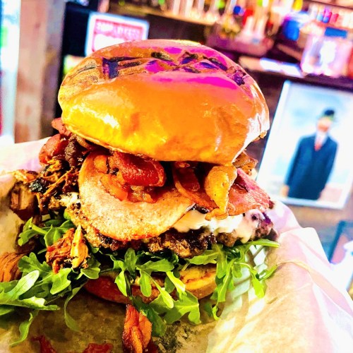 A close-up of a gourmet burger with layers of meat, bacon, lettuce, and a shiny bun, served on parchment paper.