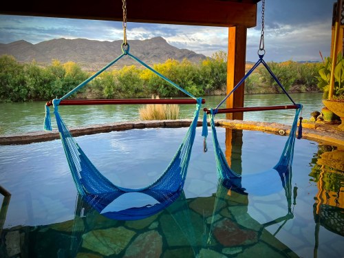 Two blue hammocks suspended over a tranquil pool, with mountains and greenery in the background under a cloudy sky.