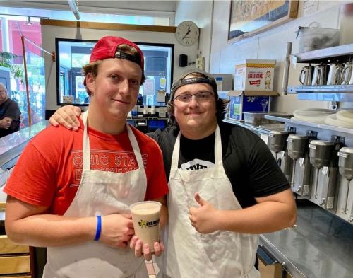 Two young men in aprons smile and pose with a cup in a cafe kitchen.