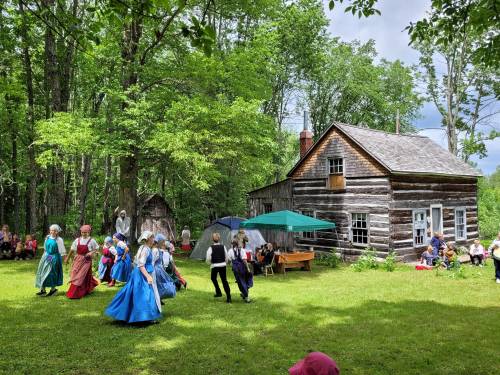 A group of people in colorful costumes dance outdoors near a log cabin, surrounded by trees and an audience.