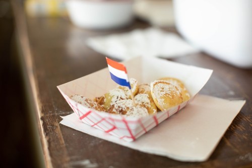 A small tray of powdered sugar-dusted pastries with a tiny Dutch flag on top, placed on a wooden table.