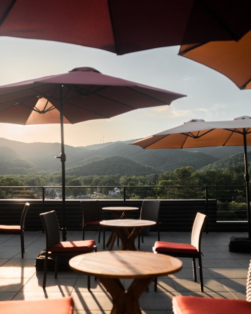 A rooftop terrace with tables and chairs under large umbrellas, overlooking mountains at sunset.