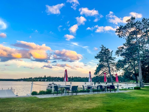 A serene lakeside scene with tables and umbrellas, surrounded by green grass and trees under a blue sky with fluffy clouds.