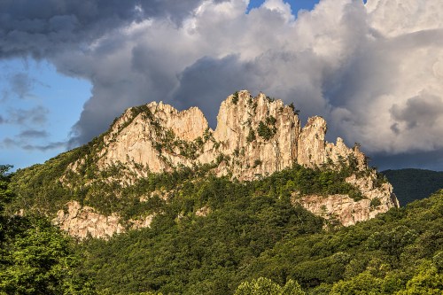 A rugged mountain peak rises above a lush green forest under a dramatic sky with clouds.