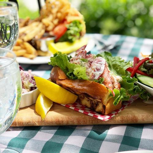 A close-up of a lobster roll with lettuce, served with lemon and a side salad on a checkered tablecloth.