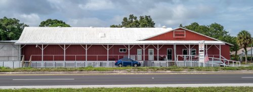 A red barn-style building with a white roof, featuring a porch and parked blue car, set against a cloudy sky.
