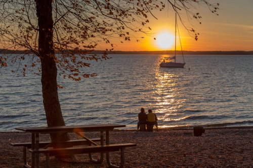 A serene sunset over a lake, with a couple sitting on the shore and a sailboat in the distance.