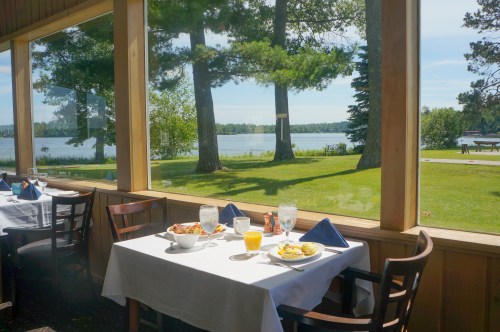 A dining table set with breakfast dishes, overlooking a serene lake and trees through large windows.