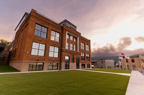 Historic brick building with large windows, set against a sunset sky and green lawn.