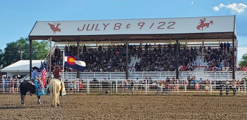 A rodeo scene with riders on horseback holding flags in front of a large crowd under a covered arena.