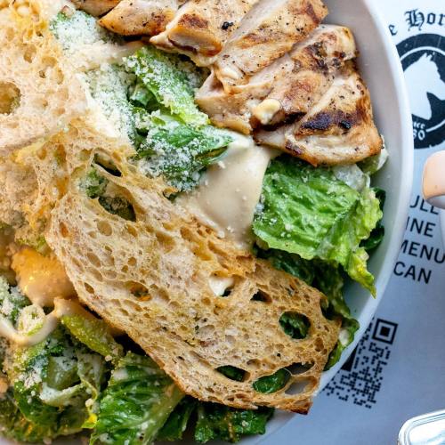 A close-up of a Caesar salad with grilled chicken, romaine lettuce, croutons, and Parmesan cheese in a white bowl.