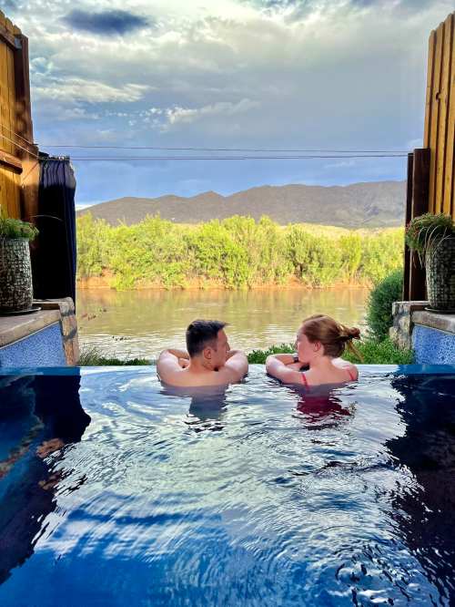 A couple relaxes in a pool, overlooking a river and mountains under a cloudy sky.